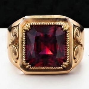Puede incluir: Un anillo de oro con una gran piedra preciosa de color rojo intenso y corte cuadrado engastada en un bisel con cuentas. La banda muestra intrincados grabados de volutas en ambos lados. El texto U.R.G. es visible en un patrón repetido.