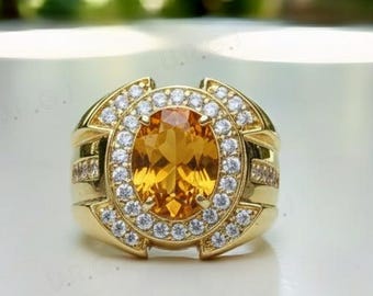 May include: A gold ring featuring an oval, orange gemstone surrounded by a halo of smaller, clear stones. The ring has a wide band with decorative elements and is set against a blurred green background.