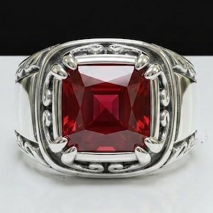 May include: A silver ring with a large, square, red gemstone. The ring has an ornate design around the gemstone and on the band. The gemstone is a deep, rich red color, and the silver band is polished.