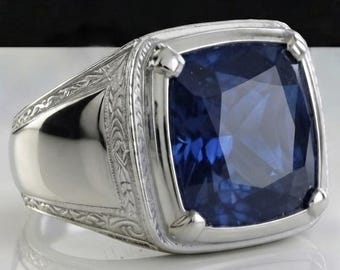 May include: A silver-toned ring featuring a large, cushion-cut deep blue gemstone set in a four-prong mounting. The band displays intricate, engraved scrollwork patterns along the sides, leading to a smooth, polished finish on the lower shank.