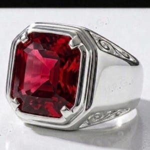 May include: A silver ring featuring a large, rectangular, deep red gemstone. The ring has a thick band and decorative detailing on the sides. The gemstone is faceted, reflecting light and creating a brilliant display.