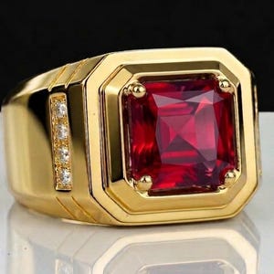 Può includere: Un anello color oro con una grande gemma rossa quadrata al centro. L'anello ha una fascia spessa ed è impreziosito da piccole pietre trasparenti su un lato. L'anello è presentato su una superficie riflettente.