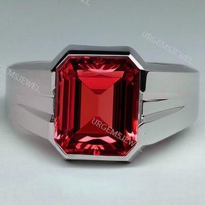 Peut inclure: Une bague en argent avec une grosse pierre rouge rectangulaire à facettes. La bague a un design moderne et géométrique avec une finition polie. Les mots "URGEMSJEWEL" sont visibles sur la bague.