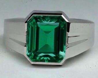 May include: A silver ring featuring a large, rectangular emerald-cut green gemstone. The ring has a wide band with a simple, modern design. The gemstone is a vibrant, deep green color, set in a polished silver setting.