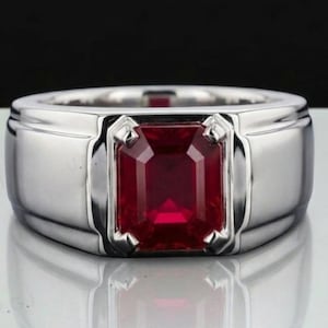 Puede incluir: Un anillo plateado con una gran gema rectangular de color rojo oscuro en el centro. El anillo tiene una banda gruesa y un acabado pulido, que refleja la luz. La gema está firmemente engastada en el diseño del anillo.