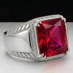 Può includere: Un anello d'argento con una grande gemma rossa vibrante di forma rettangolare. L'anello ha una fascia spessa e un bordo decorativo strutturato attorno alla gemma. L'incastonatura è in argento lucido.