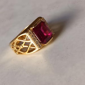 K&ouml;nnte beinhalten: Ein goldfarbener Ring mit einem gro&szlig;en, rechteckigen, rubinroten Edelstein. Der Ring hat ein dekoratives Filigranmuster am Band und ist mit kleinen, klaren Edelsteinen um den Hauptstein verziert. Ein auff&auml;lliger Ring.