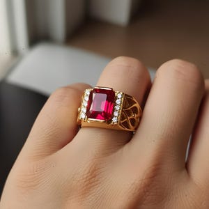 K&ouml;nnte beinhalten: Ein goldener Ring mit einem gro&szlig;en, rechteckigen, tiefroten Edelstein. Der Ring ist mit kleineren, klaren Edelsteinen und dekorativen Filigranarbeiten an den Seiten verziert. Der Ring wird an einem Finger getragen.