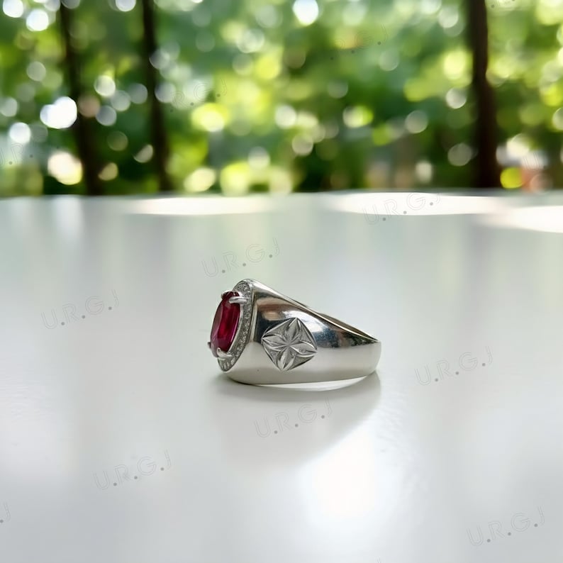 May include: A silver ring with a large oval, deep red gemstone, surrounded by smaller clear stones. The ring has a starburst design on the side. The ring is set against a blurred green background.