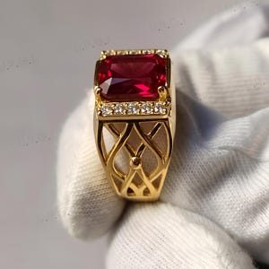 K&ouml;nnte beinhalten: Ein goldener Ring mit einem gro&szlig;en, rechteckigen, tiefroten Edelstein. Der Ring ist mit kleinen, klaren Steinen verziert und hat ein offenes, dekoratives Filigranmuster am Band.