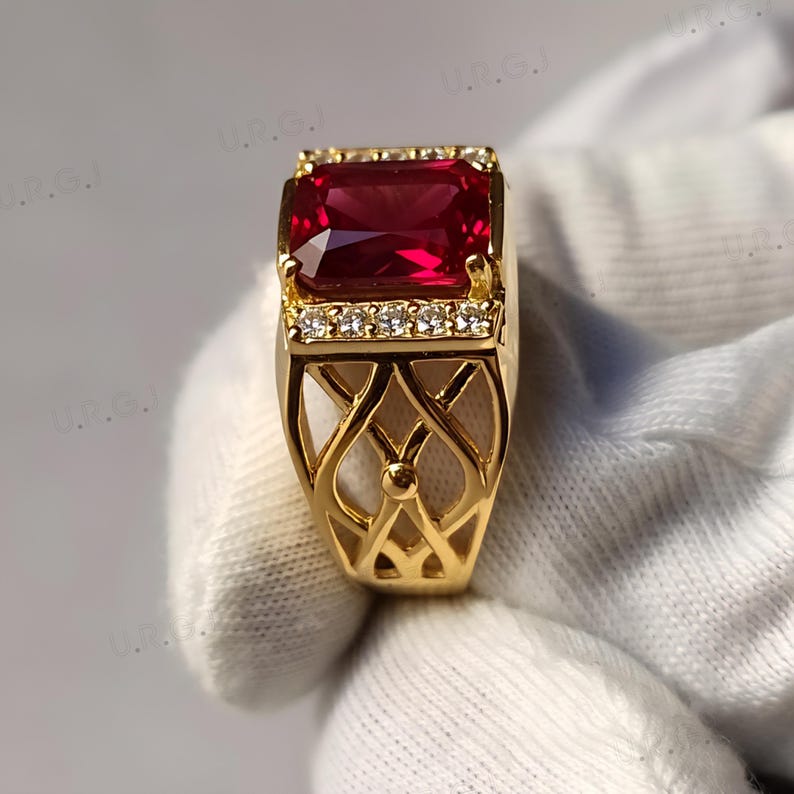 K&ouml;nnte beinhalten: Ein goldener Ring mit einem gro&szlig;en, rechteckigen, tiefroten Edelstein. Der Ring ist mit kleinen, klaren Steinen verziert und hat ein offenes, dekoratives Filigranmuster am Band.