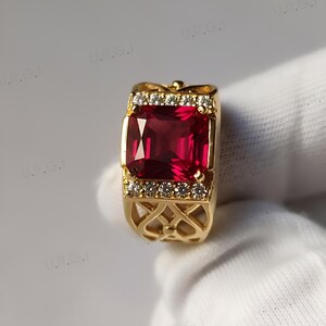K&ouml;nnte beinhalten: Ein goldener Ring mit einem gro&szlig;en, rechteckigen, tiefroten Edelstein. Der Ring ist mit kleinen, klaren Edelsteinen und aufwendigen Filigranarbeiten am Band verziert. Ein auff&auml;lliges Schmuckst&uuml;ck.