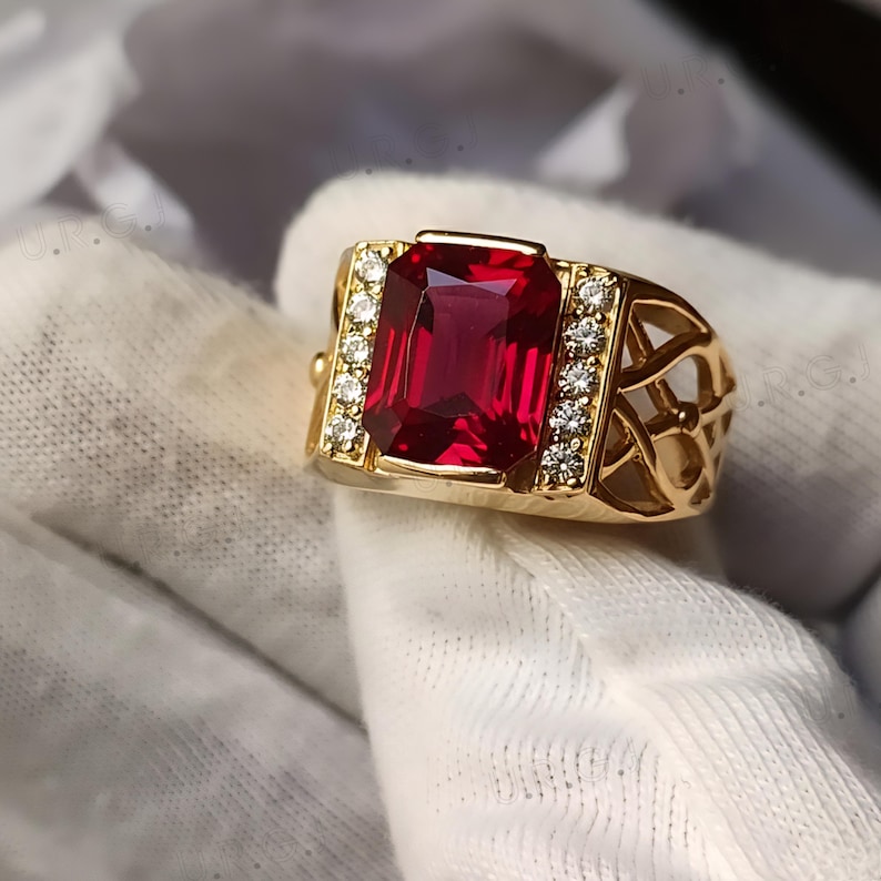 K&ouml;nnte beinhalten: Ein goldener Ring mit einem gro&szlig;en, rechteckigen, tiefroten Edelstein. Der Ring ist mit kleinen, klaren Edelsteinen auf beiden Seiten des zentralen Steins verziert. Das Band hat ein offenes, dekoratives Design.