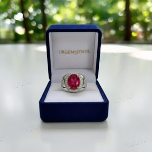 May include: A silver ring with a large oval ruby gemstone, surrounded by smaller clear stones. The ring is displayed in a blue velvet box with the text "URGEMSJEWEL" on the lid.