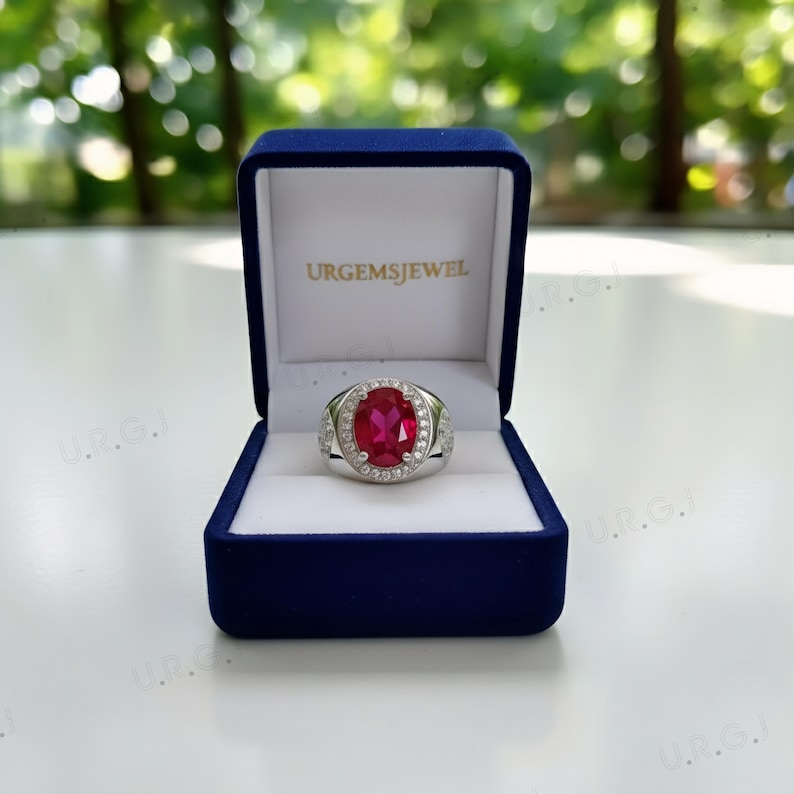 May include: A silver ring with a large oval ruby gemstone, surrounded by smaller clear stones. The ring is displayed in a blue velvet box with the text "URGEMSJEWEL" on the lid.