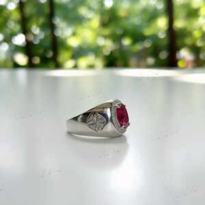 May include: A silver ring with a large oval-cut, deep red gemstone. The gemstone is surrounded by a halo of smaller, clear stones. The ring has a decorative starburst design on the side.