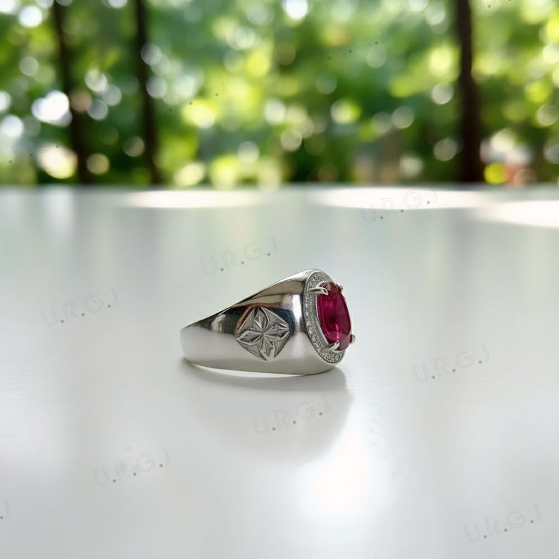 May include: A silver ring with a large oval-cut, deep red gemstone. The gemstone is surrounded by a halo of smaller, clear stones. The ring has a decorative starburst design on the side.
