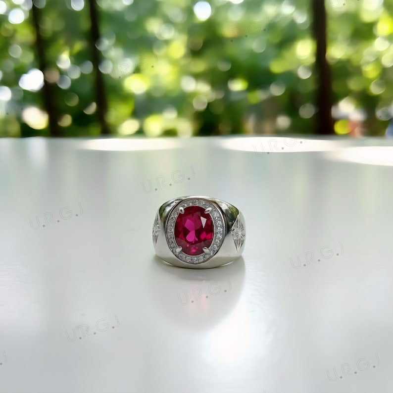 May include: A silver ring with a large, oval-cut, vibrant red gemstone as the centerpiece. The gemstone is surrounded by a halo of smaller, clear stones. The ring has a wide band with decorative starburst patterns on the sides.