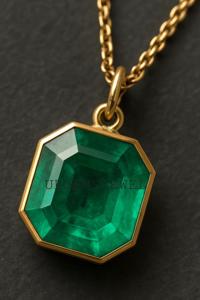 May include: A gold necklace with a large, emerald-cut green gemstone pendant. The pendant is set in a gold frame and hangs from a delicate gold chain. The text "U GEMS JEWEL" is visible on the pendant.