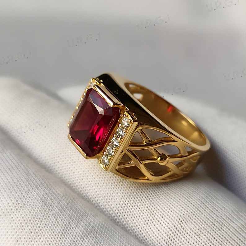 K&ouml;nnte beinhalten: Ein goldfarbener Ring mit einem gro&szlig;en, rechteckigen, tiefroten Edelstein als Mittelpunkt. Der Ring ist mit kleinen, klaren Akzentsteinen verziert und hat ein filigranes Design am Band. Der Ring liegt auf einer wei&szlig;en Oberfl&auml;che.
