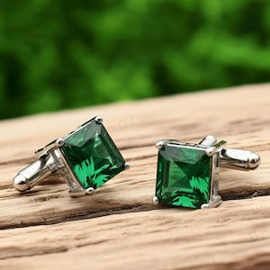 May include: A pair of square-cut, emerald-green cufflinks with a silver-tone metal setting. The cufflinks are presented on a wooden surface with a blurred green backdrop. The cufflinks have a classic design.