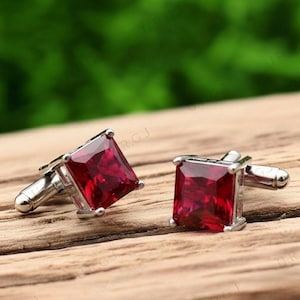 Puede incluir: Un par de gemelos cuadrados con un centro de piedra preciosa rojo vibrante. Las piedras preciosas están engastadas en un marco de metal plateado. Los gemelos se exhiben sobre una superficie de madera.
