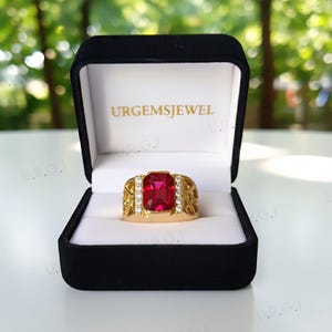 K&ouml;nnte beinhalten: Ein goldfarbener Ring mit einem gro&szlig;en, rechteckigen roten Edelstein und kleinen wei&szlig;en Edelsteinen. Der Ring wird in einer schwarzen Schatulle mit dem Text "URGEMSJEWEL" pr&auml;sentiert.