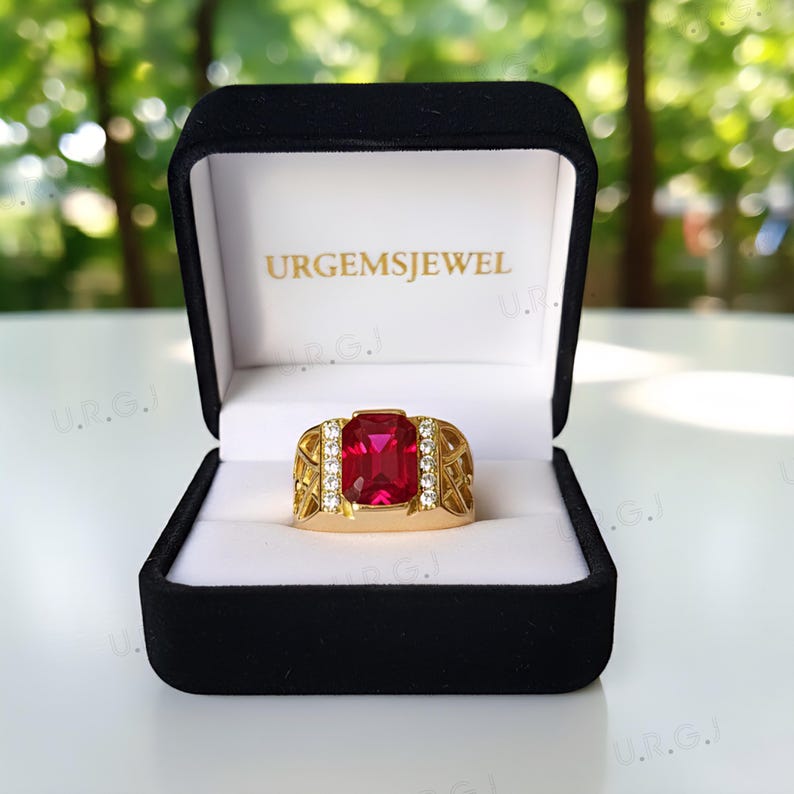 K&ouml;nnte beinhalten: Ein goldfarbener Ring mit einem gro&szlig;en, rechteckigen roten Edelstein und kleinen wei&szlig;en Edelsteinen. Der Ring wird in einer schwarzen Schatulle mit dem Text "URGEMSJEWEL" pr&auml;sentiert.
