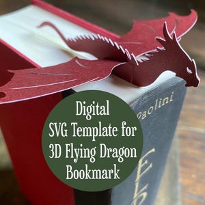 Pode incluir: Um marcador de dragão 3D vermelho inserido em um livro. O dragão de papel tem asas abertas. O livro tem páginas vermelhas e brancas. O texto na imagem diz "Digital SVG Template for 3D Flying Dragon Bookmark."