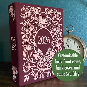 Op de afbeelding: Een bordeauxrode boekomslag met een bloemenontwerp en het jaar 2026. De omslag heeft een decoratief bloemenontwerp in een lichtere roze tint. De tekst "Customizable book front cover, back cover, and spine SVG files" staat in een groene cirkel.