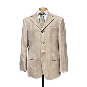 May include: A beige blazer with a subtle vertical pinstripe pattern, featuring a three-button front closure, notched lapels, two flap pockets, and a single chest pocket. The blazer is worn over a white collared shirt with a green patterned tie.