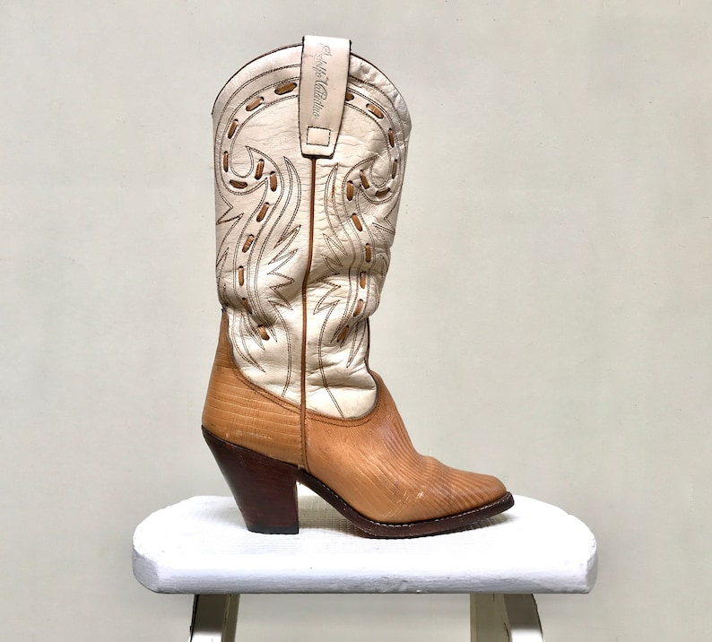 May include: A vintage cowboy boot with a cream-colored upper featuring decorative stitching and a tan leather foot. The boot has a high heel and is displayed on a white stool. The text "R.M. Williams" is visible on the boot's upper.