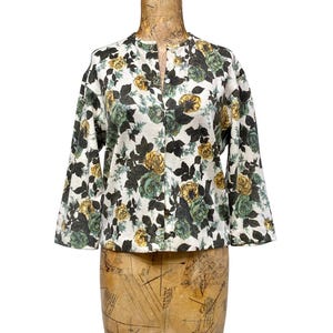 May include: A white long-sleeve button-down shirt featuring a floral pattern with yellow and dark green roses and dark green leaves. The shirt has a round neckline and a straight hem, displayed on a vintage-style dress form mannequin.