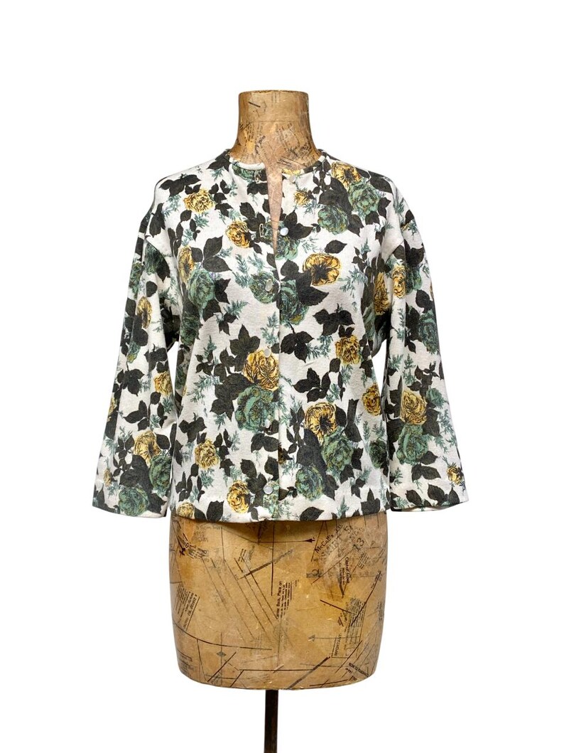May include: A white long-sleeve button-down shirt featuring a floral pattern with yellow and dark green roses and dark green leaves. The shirt has a round neckline and a straight hem, displayed on a vintage-style dress form mannequin.