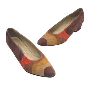 May include: Two suede low-heeled shoes in shades of brown, orange, and tan. The shoes have a pointed toe and a low heel. The design features curved color blocks. The shoes are set against a white background.