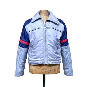 May include: A vintage light blue jacket with a zip-up front, featuring navy blue and red accents on the sleeves. The jacket is displayed on a wooden mannequin, showcasing its retro design and style.