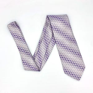 May include: A patterned necktie in shades of purple and gray, arranged on a white surface. The tie features a repeating geometric design of small squares, creating a textured appearance. The tie is a classic accessory for formal or business attire.