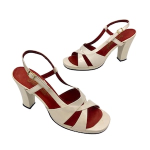 May include: Pair of cream-colored vintage high-heeled sandals with red insoles. The sandals feature a criss-cross design across the toes, ankle straps, and chunky heels. The heels are approximately 3 inches (7.6 cm) high.