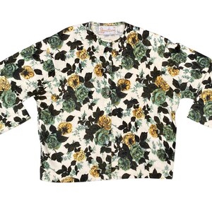May include: A long-sleeved button-down cardigan featuring a cream-colored base with a dense floral print of dark green, muted teal, and golden yellow roses and leaves. The garment includes a round neckline and a vertical row of buttons.