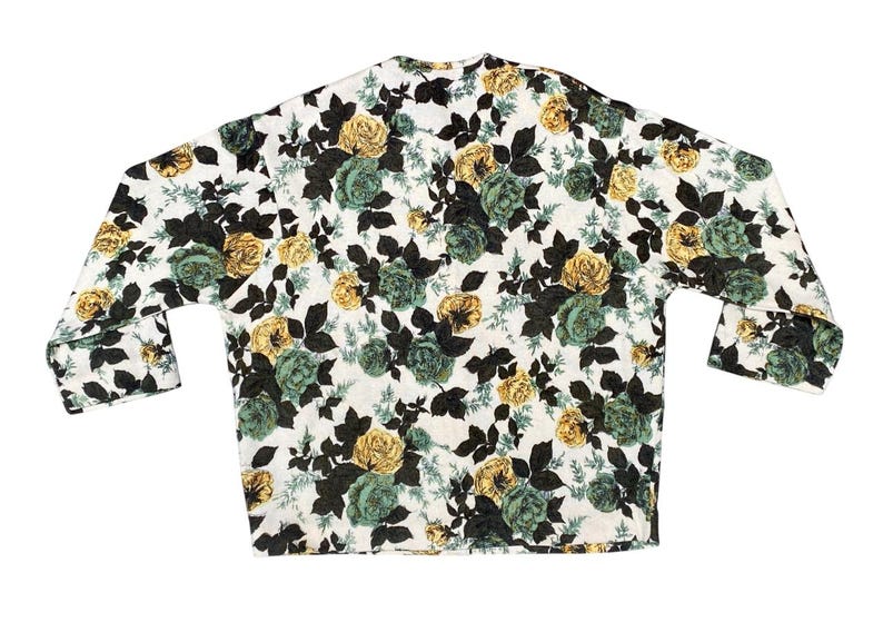 May include: A long-sleeved shirt featuring a white base with a dense floral pattern of yellow and teal roses, accompanied by dark brown leaves and stems. The garment has a straight hemline and a simple neckline, showcasing a repeating botanical print.