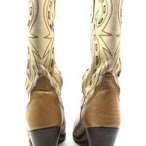 May include: A pair of vintage cowboy boots. The boots are cream-colored with tan accents and dark brown heels. The shafts feature decorative stitching and cut-out designs. The heels measure approximately 3 inches.