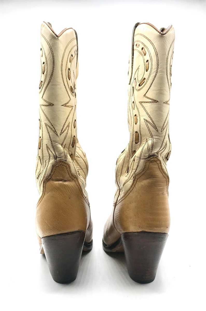 May include: A pair of vintage cowboy boots. The boots are cream-colored with tan accents and dark brown heels. The shafts feature decorative stitching and cut-out designs. The heels measure approximately 3 inches.