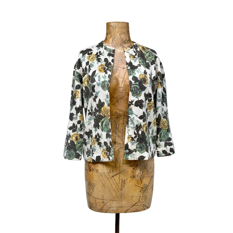 May include: A white open-front jacket featuring a floral pattern with dark green, light green, and yellow roses. The garment has three-quarter length sleeves and a round neckline, displaying a consistent botanical print across the entire fabric surface.