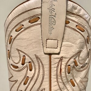 May include: Close-up of a cream-colored cowboy boot with decorative stitching and a pull strap. The boot features brown stitching and accents, with the brand name "Adolfo Valentino" on the pull strap. The boot's design includes cut-out details.