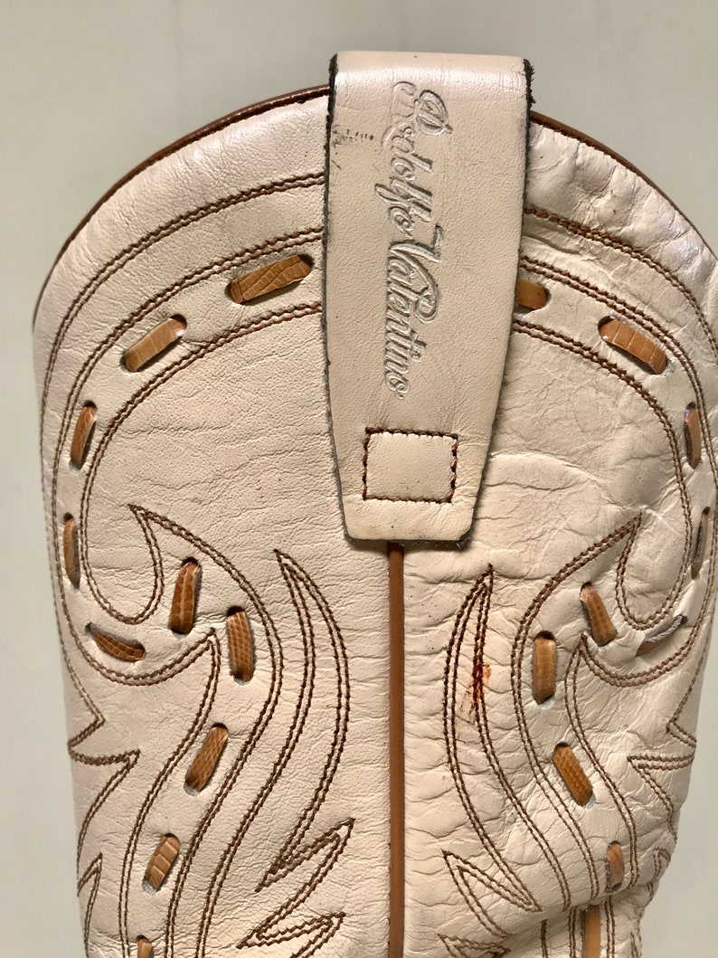 May include: Close-up of a cream-colored cowboy boot with decorative stitching and a pull strap. The boot features brown stitching and accents, with the brand name "Adolfo Valentino" on the pull strap. The boot's design includes cut-out details.
