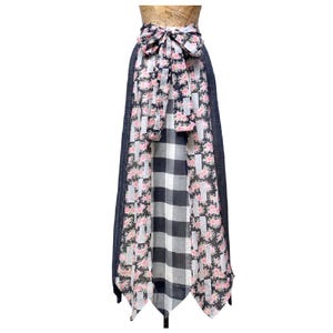 May include: A long skirt with a bow at the waist. The skirt features a mix of patterns, including a floral print, a black and white checkered pattern, and a dark blue denim-like fabric. The bow is made of the floral print fabric.