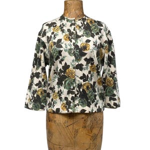 May include: A long-sleeved button-down jacket featuring a cream-colored base with a dense floral pattern of yellow and dark green roses and dark green leaves. The garment has a round neckline and a straight hem, displaying a consistent botanical print.