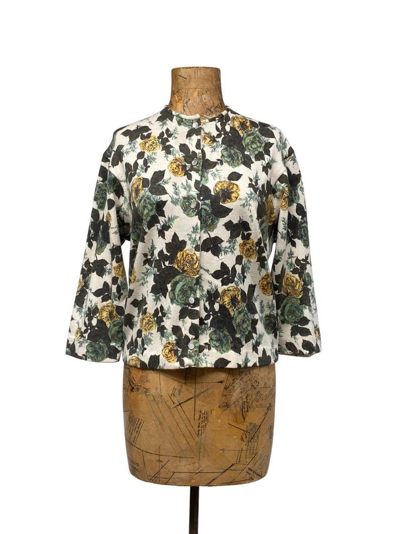 May include: A long-sleeved button-down jacket featuring a cream-colored base with a dense floral pattern of yellow and dark green roses and dark green leaves. The garment has a round neckline and a straight hem, displaying a consistent botanical print.