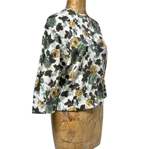 May include: A long-sleeve button-down shirt featuring a white base with a repeating floral pattern in shades of dark green, muted yellow, and black. The garment has a round neckline and a front button closure with small white buttons.