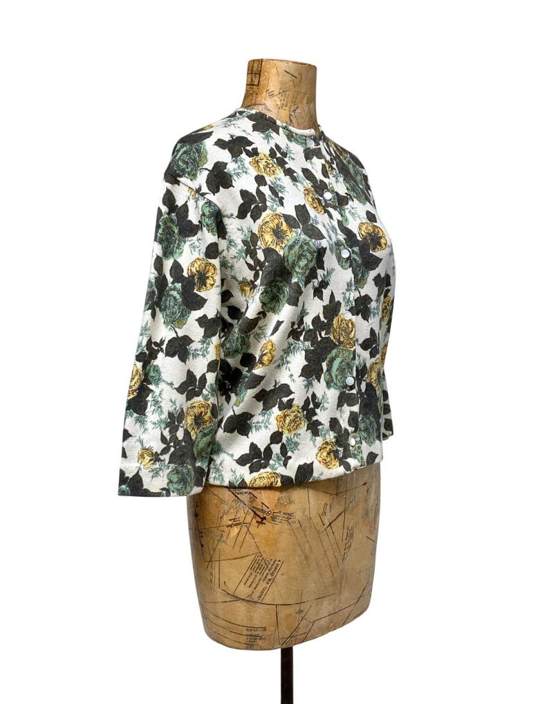 May include: A long-sleeve button-down shirt featuring a white base with a repeating floral pattern in shades of dark green, muted yellow, and black. The garment has a round neckline and a front button closure with small white buttons.